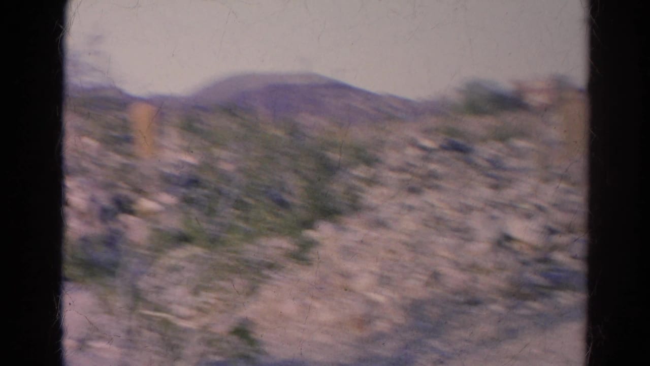 Motion-blurred 8mm view across the Silver Lake hillside, with rocks, scrub vegetation, a wooden marker-like post, and distant low ridges.