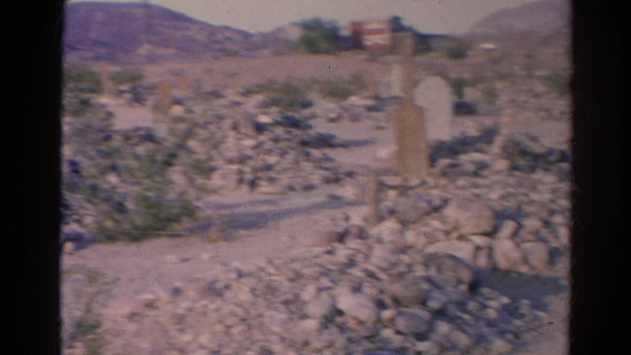 1966 Silver Lake home-movie frame showing rock piles, pale posts, scrub vegetation, distant hills, and a small background structure.