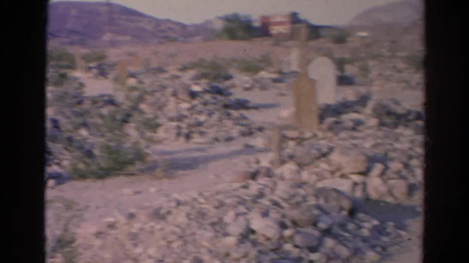 1966 Silver Lake home-movie frame showing rock piles, pale posts, scrub vegetation, distant hills, and a small background structure.