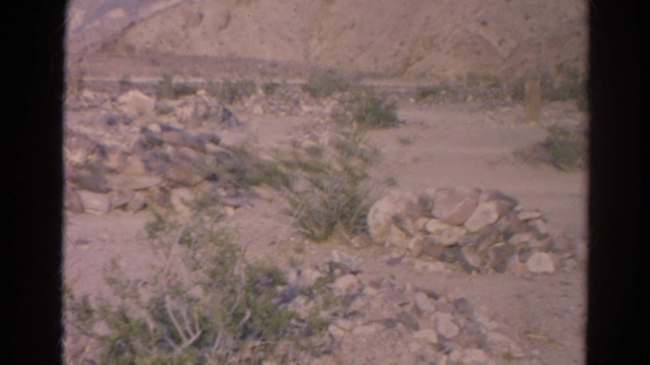 1966 Silver Lake frame with a low stone pile in the foreground, scattered desert plants, and a pale rocky hillside in the distance.