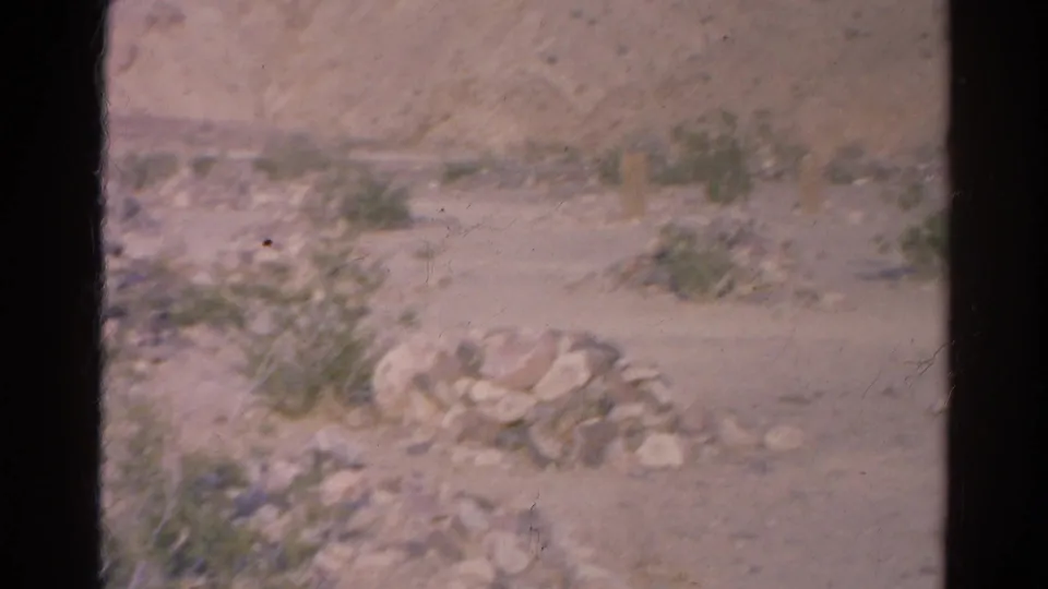 1966 8mm frame of dry desert terrain in the Silver Lake, California area, with rocks, shrubs and a low hillside.