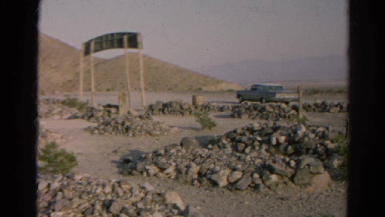 1966 Silver Lake, California desert roadside view with a dark sign on posts, rock borders, hills and a parked car.