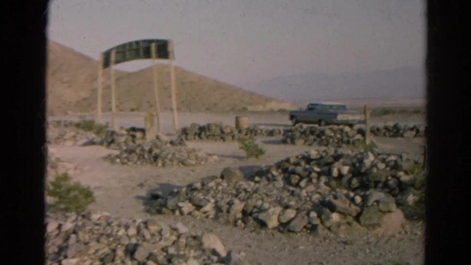 1966 Silver Lake, California desert roadside view with a dark sign on posts, rock borders, hills and a parked car.