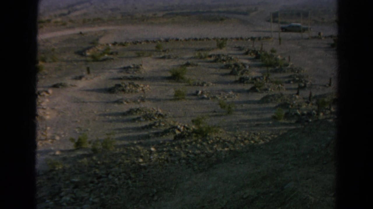 Late-exposure 1966 8mm frame of Silver Lake desert ground, rock borders, sparse shrubs and the roadside sign partly visible.