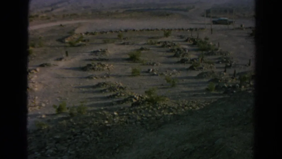 Late-exposure 1966 8mm frame of Silver Lake desert ground, rock borders, sparse shrubs and the roadside sign partly visible.