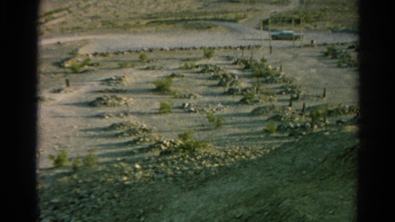 1966 8mm view of an arid Silver Lake, California burial ground with simple markers, dry terrain, sparse vegetation, and a car near the road.