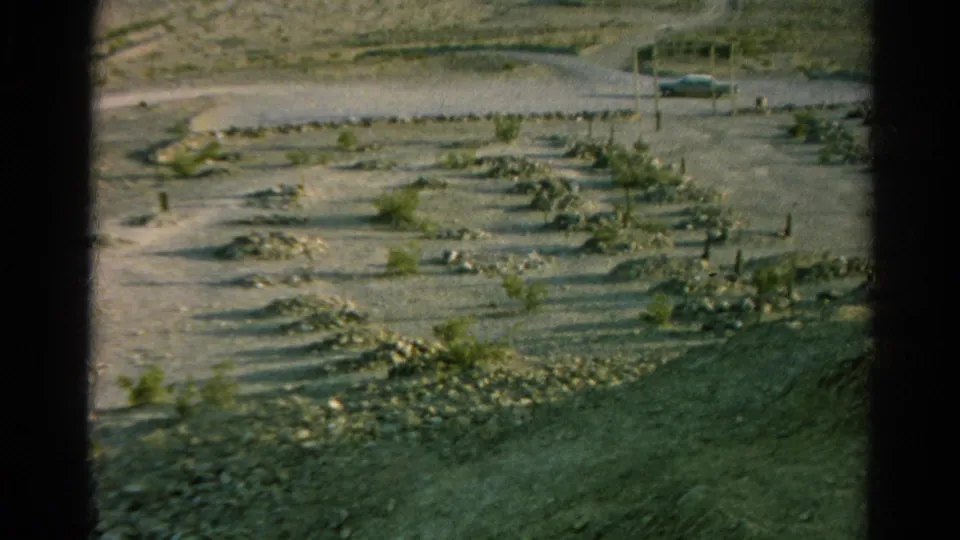 1966 8mm view of an arid Silver Lake, California burial ground with simple markers, dry terrain, sparse vegetation, and a car near the road.