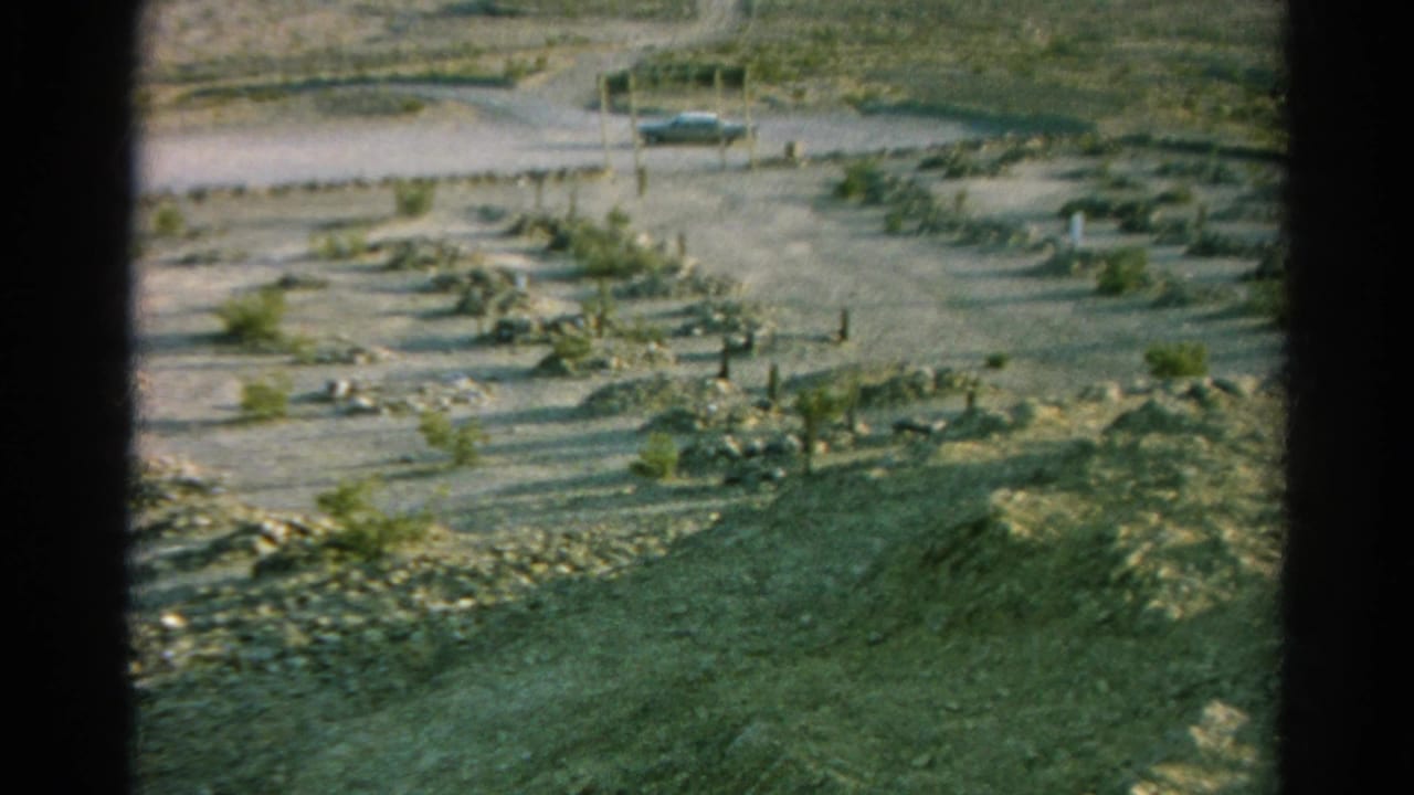 Home-movie frame from Silver Lake, California showing a barren burial ground with simple posts, dry plants, and a parked car near the road.