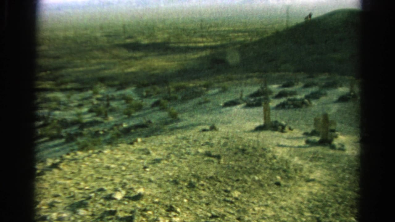 1966 8mm frame of a dry Silver Lake, California landscape with rocky ground, sparse vegetation, upright forms, and a distant hill.
