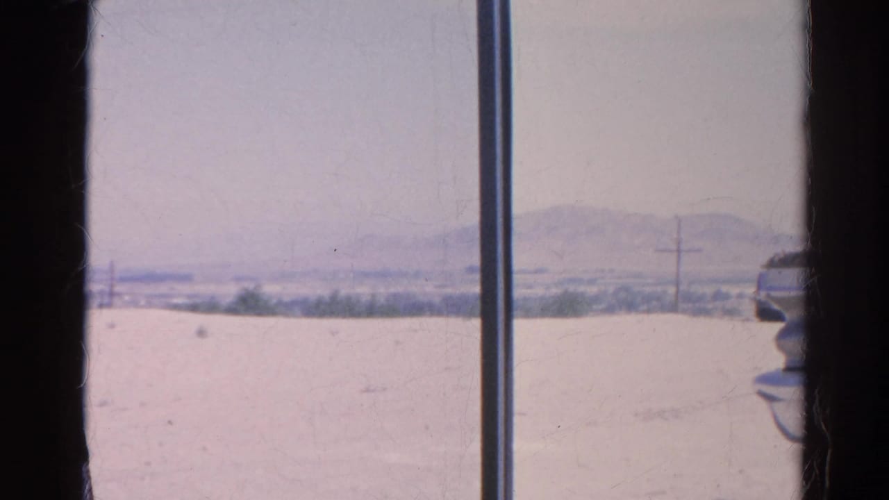 A 1966 8mm frame appears shot through a vehicle window, looking toward pale open ground, utility poles, distant hills, and part of a car.