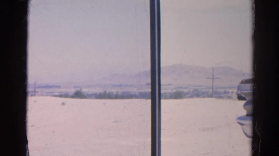 A 1966 8mm frame appears shot through a vehicle window, looking toward pale open ground, utility poles, distant hills, and part of a car.