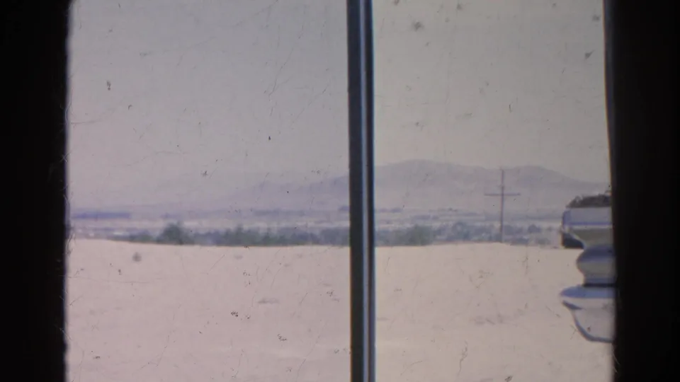 Window-framed 1966 California view with dry foreground, distant low mountains, utility pole, and the partial edge of a parked vehicle.