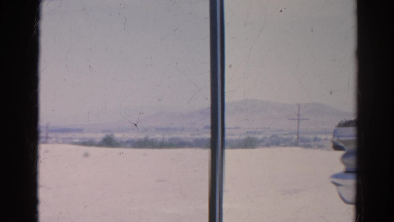 Faded 8mm frame of the Silver Lake area with dry open ground, low hills, sparse vegetation, and a utility pole seen from indoors.