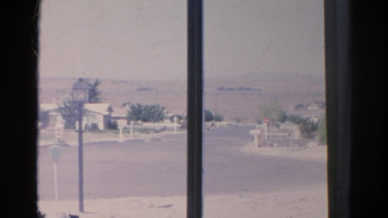 Window-divided view of a quiet street with mailboxes, a lamppost, low houses, shrubs, and hazy hills.