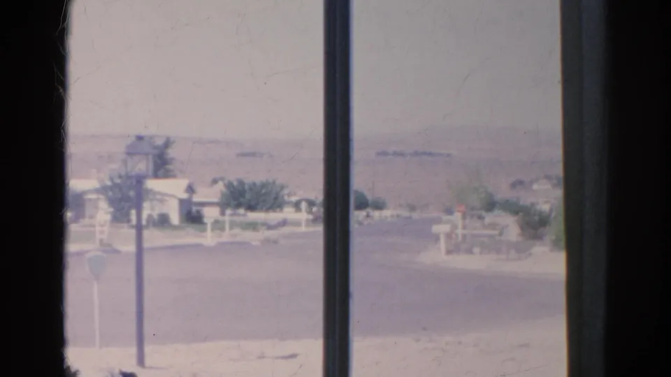 1966 Silver Lake home-movie frame looking through divided window glass at a quiet street, mailboxes, a lamppost, low homes, and haze.