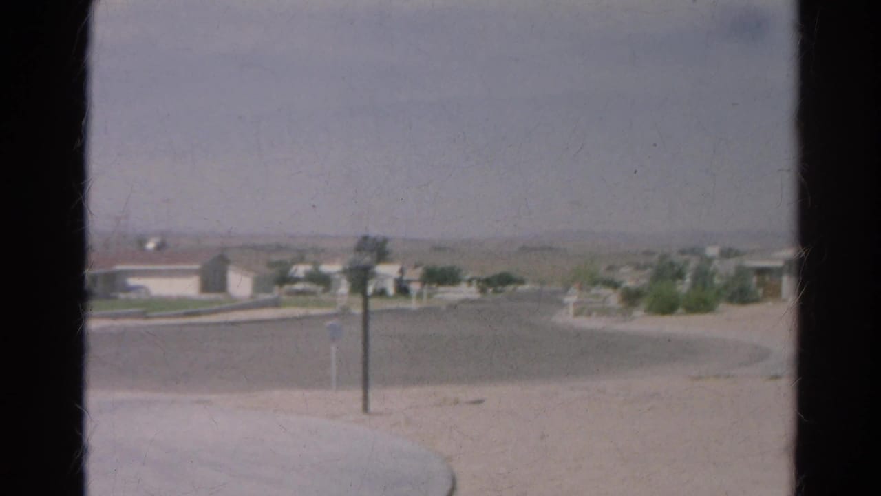 1966 8mm Silver Lake view from inside a home toward a curved road, pale sandy ground, mailboxes, a lamppost, and distant houses.
