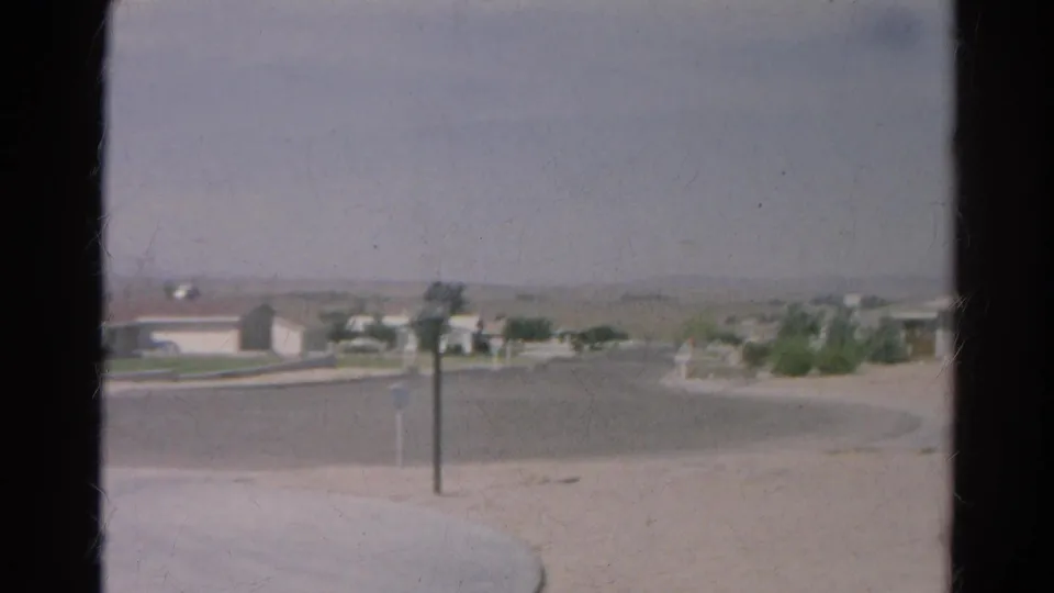 1966 8mm Silver Lake view from inside a home toward a curved road, pale sandy ground, mailboxes, a lamppost, and distant houses.
