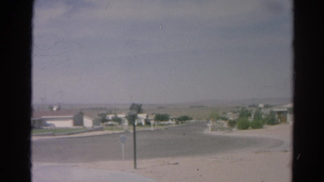 1966 Silver Lake 8mm frame through window glass showing a quiet curved road, low homes, sparse shrubs, mailboxes, and a hazy horizon.