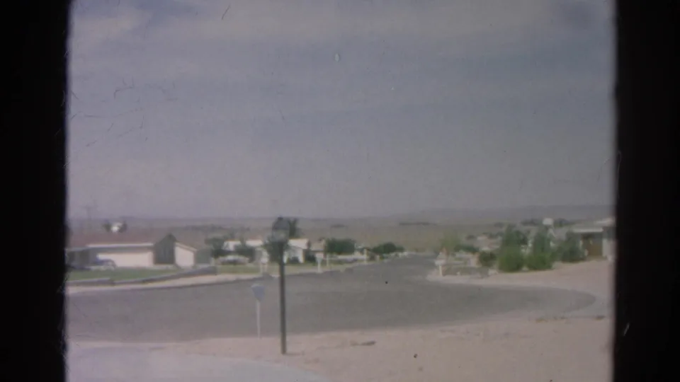 1966 Silver Lake 8mm frame through window glass showing a quiet curved road, low homes, sparse shrubs, mailboxes, and a hazy horizon.