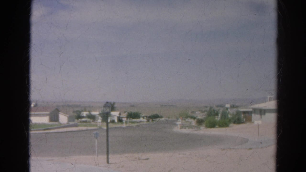Residential cul-de-sac with low houses, posts, curb line, and hazy background hills.