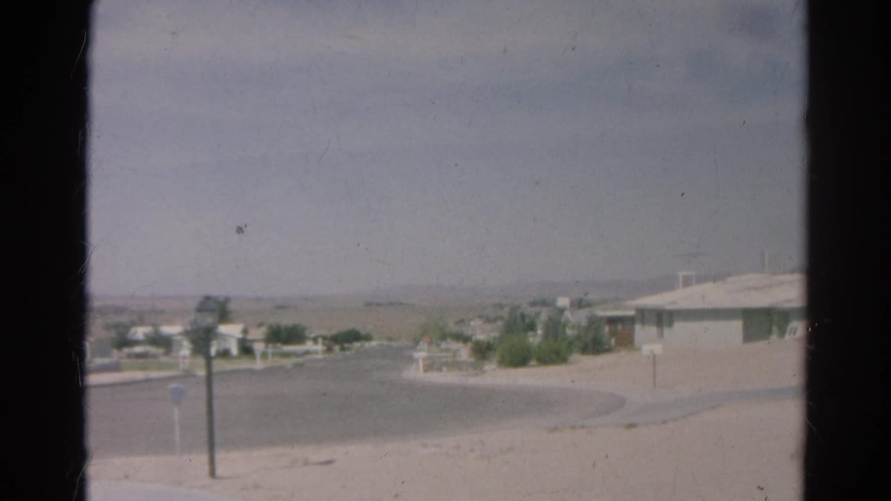 1966 Silver Lake home-movie frame showing a quiet cul-de-sac, low houses, arid ground, and a hazy sky.