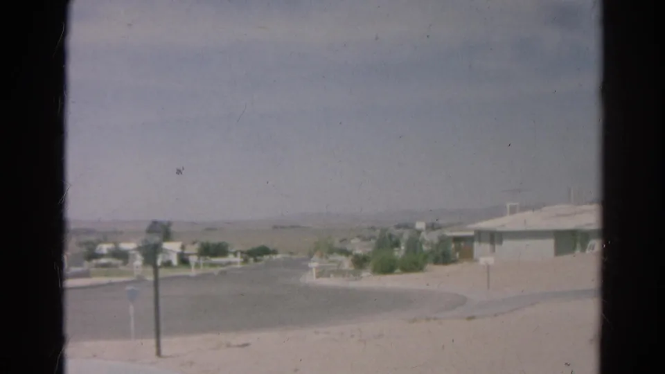 1966 Silver Lake home-movie frame showing a quiet cul-de-sac, low houses, arid ground, and a hazy sky.
