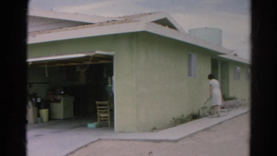 1966 Silver Lake frame showing a woman in a white dress beside a pale green house near an open garage and side walkway.
