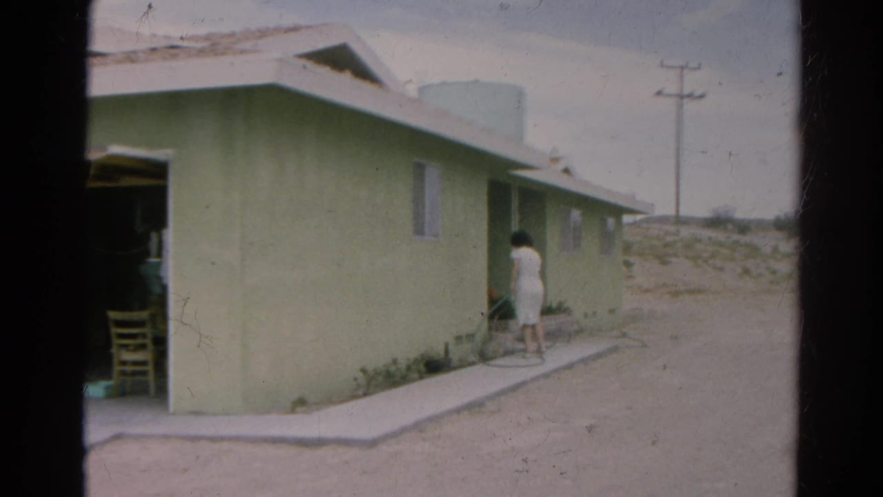 Silver Lake home-movie frame with a woman beside a pale green house, open garage edge, dirt lot, and utility pole beyond.