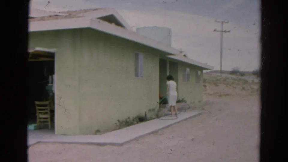 Silver Lake home-movie frame with a woman beside a pale green house, open garage edge, dirt lot, and utility pole beyond.