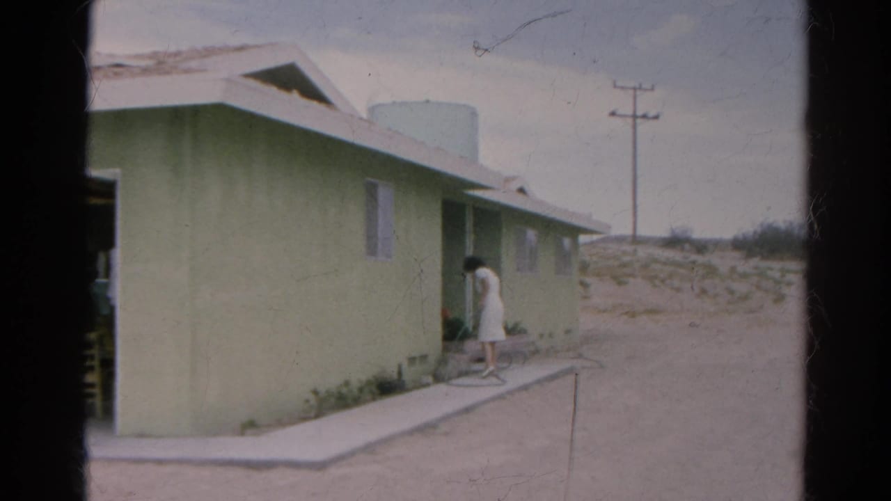 1966 Silver Lake home-movie frame showing a woman in a white dress by a pale green house, with a roof tank and utility pole in the background.