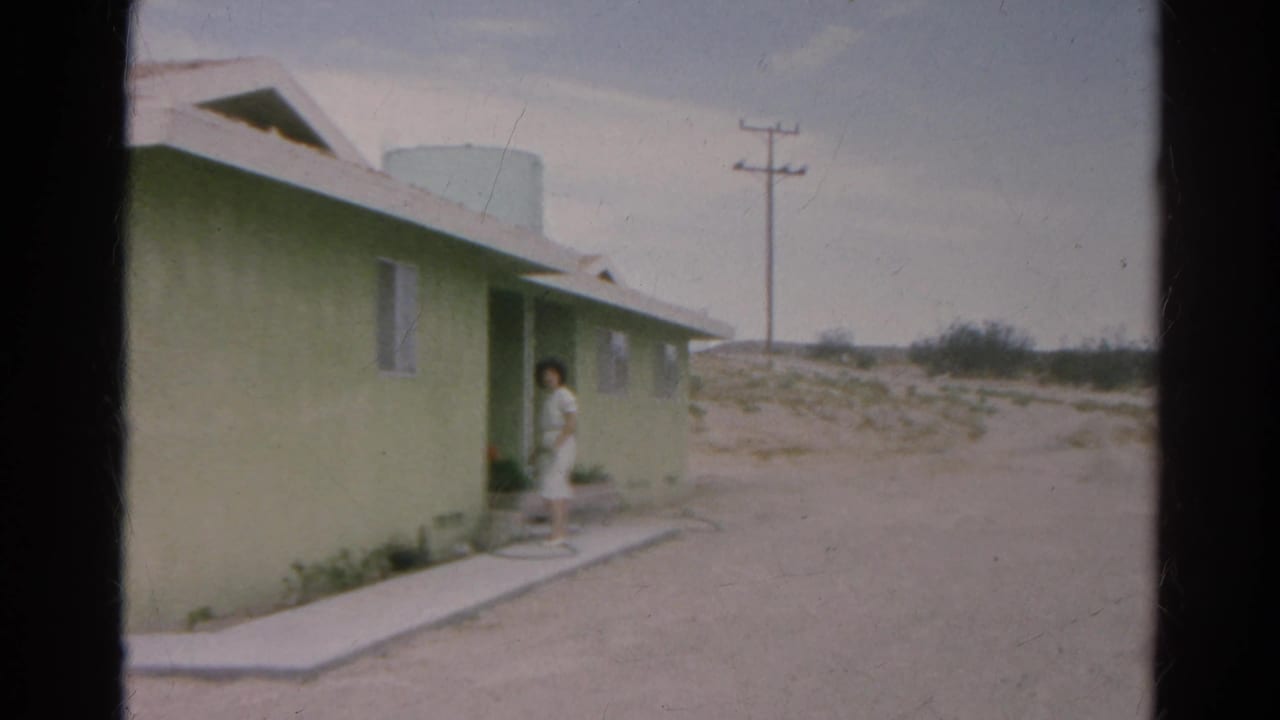 1966 Silver Lake frame with a woman in a white dress at the entrance of a pale green single-story home in a dry landscape.