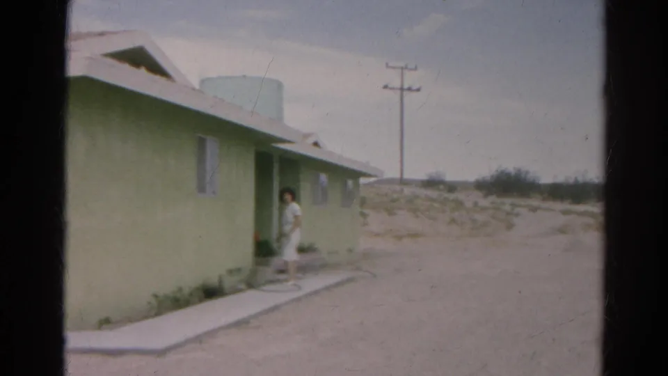 1966 Silver Lake frame with a woman in a white dress at the entrance of a pale green single-story home in a dry landscape.