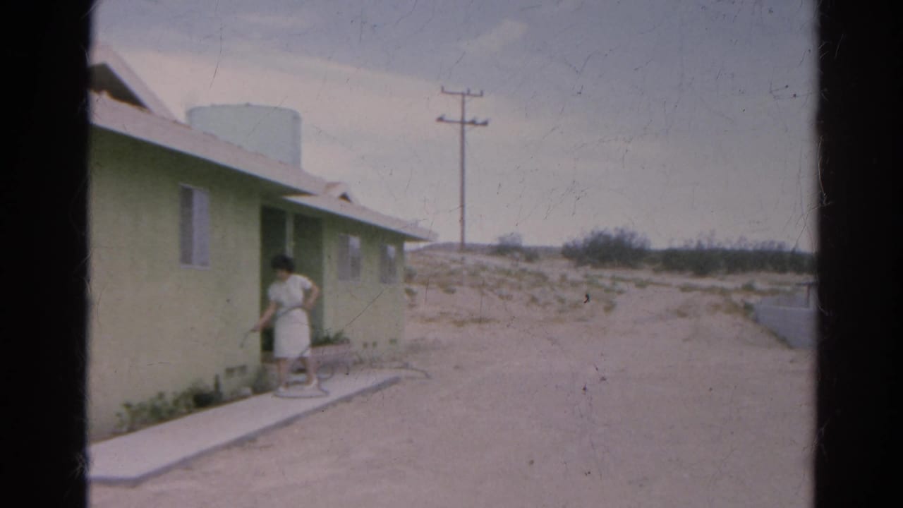 1966 Silver Lake home-movie frame showing a woman tending plants or hose beside a pale green house and sandy open ground.