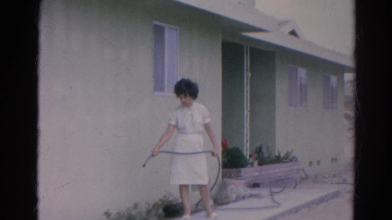 Closer 1966 Silver Lake frame of a woman in a white dress watering plants with a hose beside a pale green home exterior.