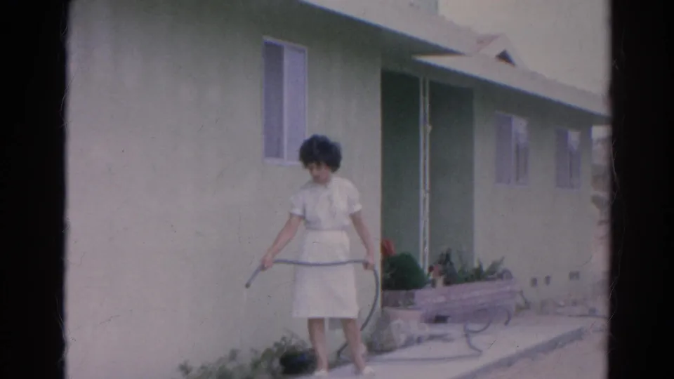 Closer 1966 Silver Lake frame of a woman in a white dress watering plants with a hose beside a pale green home exterior.