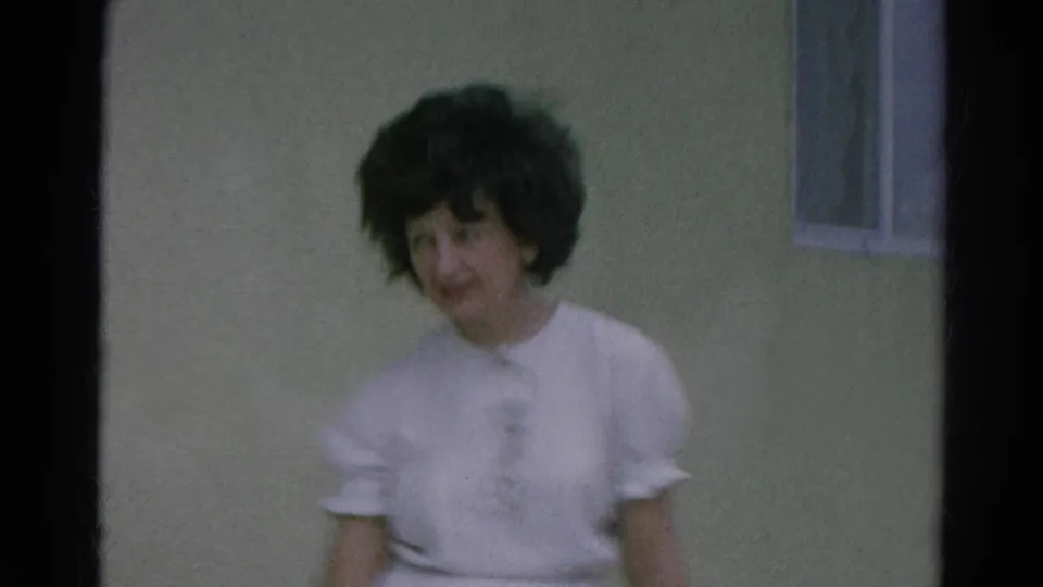 The woman faces the camera near the green stucco wall and window in the Silver Lake home-movie scene.