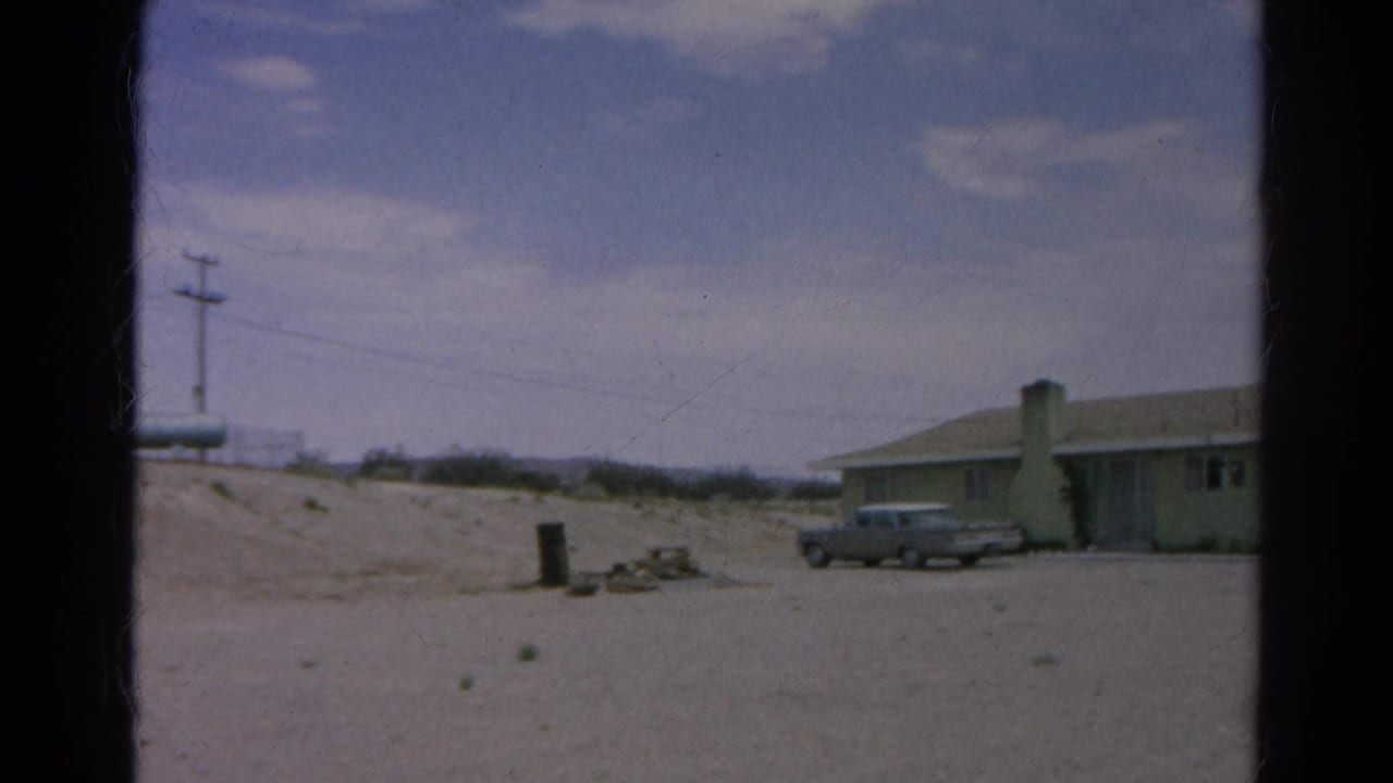 1966 8mm frame from Silver Lake, California, showing a low pale green house, chimney, parked silver sedan, sandy ground, and utility pole.