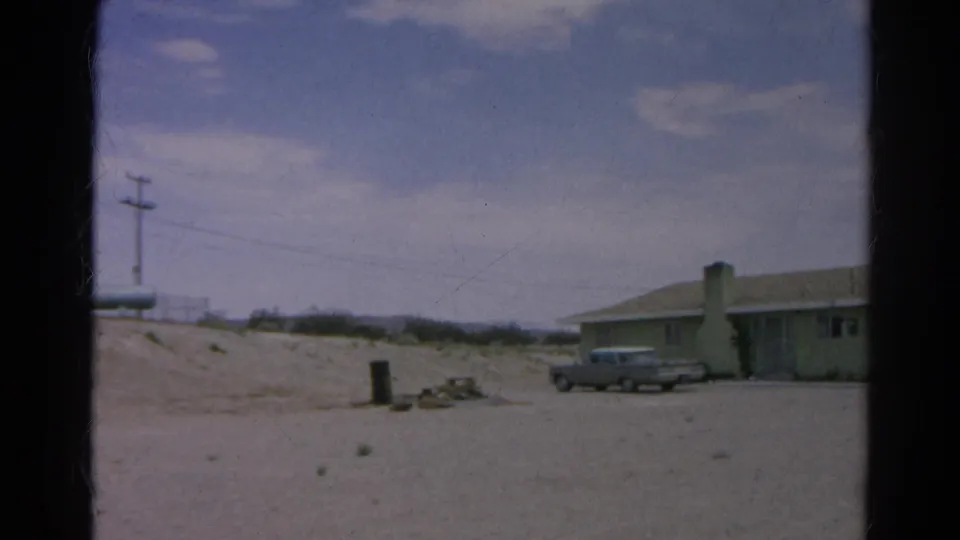 1966 8mm frame from Silver Lake, California, showing a low pale green house, chimney, parked silver sedan, sandy ground, and utility pole.