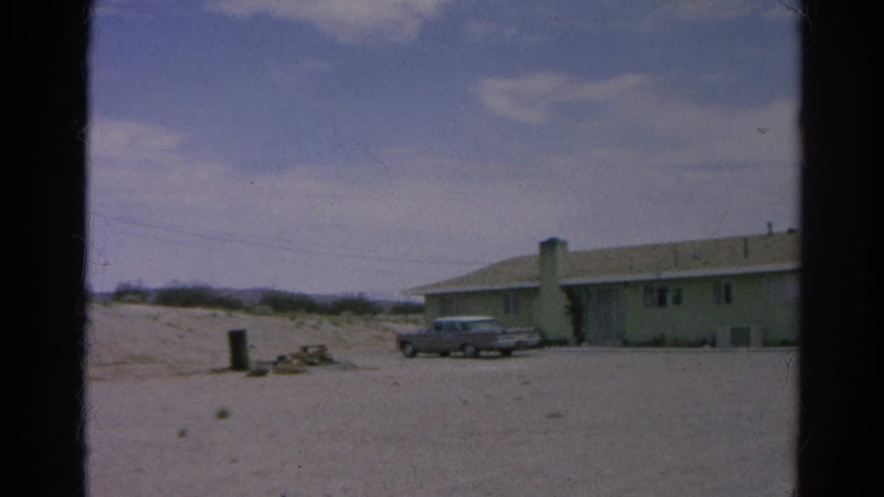 1966 Silver Lake home-movie frame showing the house and silver sedan farther right in the pan, with sandy foreground and low hills beyond.