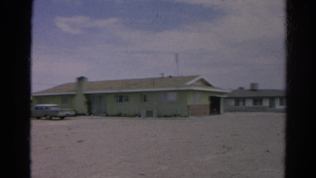 1966 8mm frame from Silver Lake, California showing a pale low-rise roadside building, gravel foreground, and a parked silver sedan.