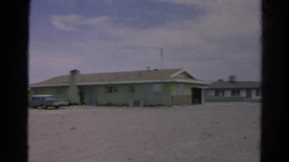 1966 8mm frame from Silver Lake, California showing a pale low-rise roadside building, gravel foreground, and a parked silver sedan.