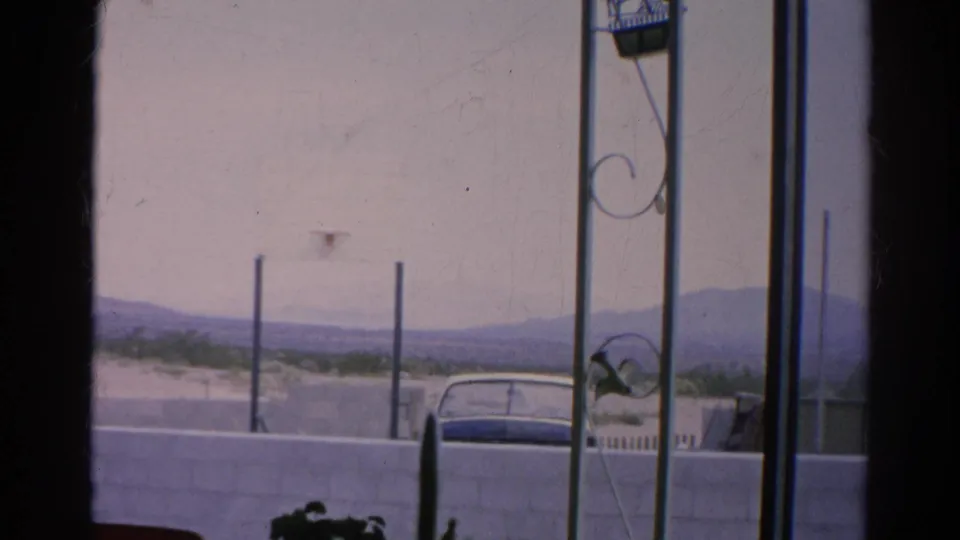 1966 Silver Lake home-movie view of a parked car behind a white block wall, framed by porch metalwork and distant hills.