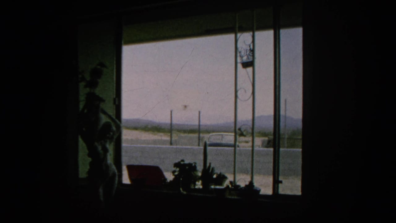 1966 Silver Lake home-movie frame from inside a dark room, with a window view of a parked car, road, fence posts, and hazy hills.
