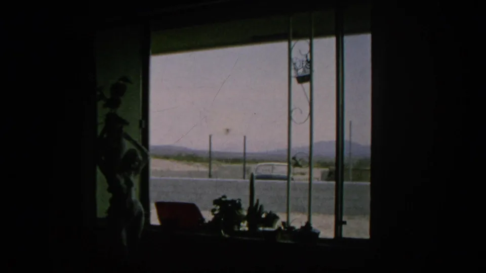 1966 Silver Lake home-movie frame from inside a dark room, with a window view of a parked car, road, fence posts, and hazy hills.