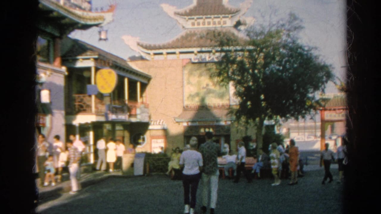 Outdoor 1966 California home-movie frame showing visitors near Chinese-style architecture, with a tree, parked cars, and a small open plaza.