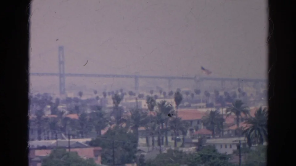 Hazy 1966 Silver Lake-area view of palms, rooftops, and a distant bridge-like span, bordered by dark 8mm frame edges.