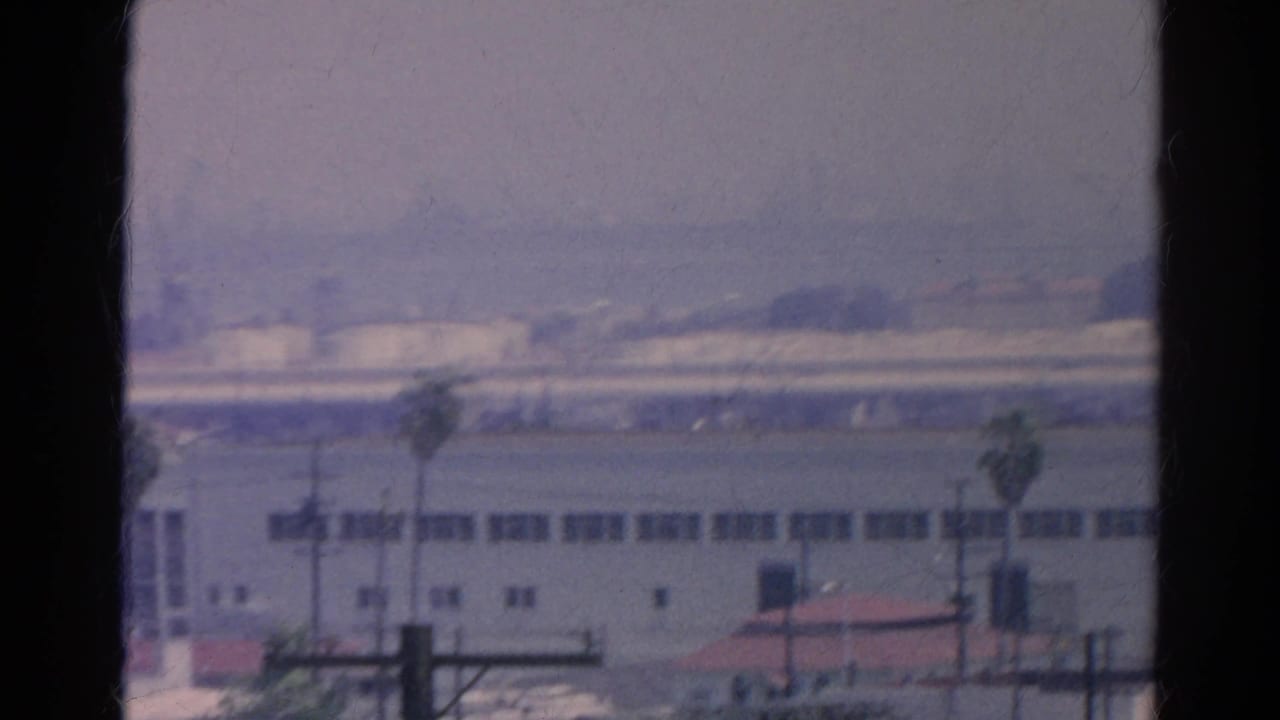 Distant industrial buildings and palm trees seen through dark window framing.