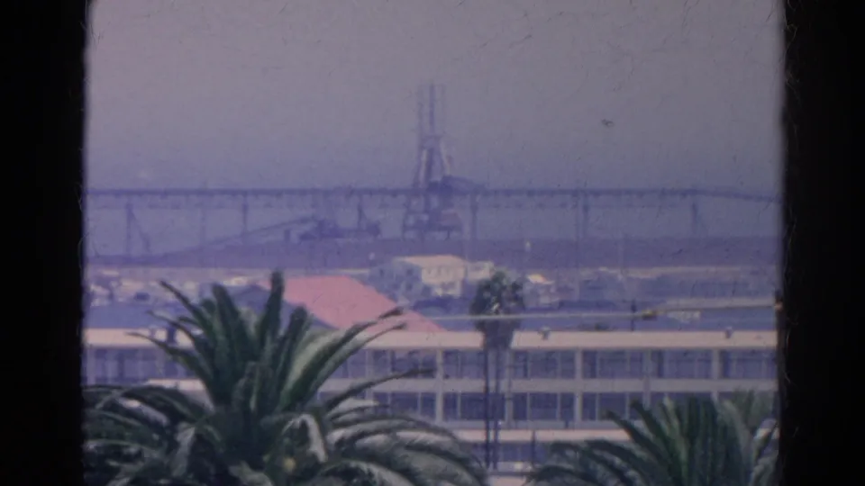 1966 California 8mm frame showing palm fronds, waterfront buildings, a long bridge, and a crane through a window.