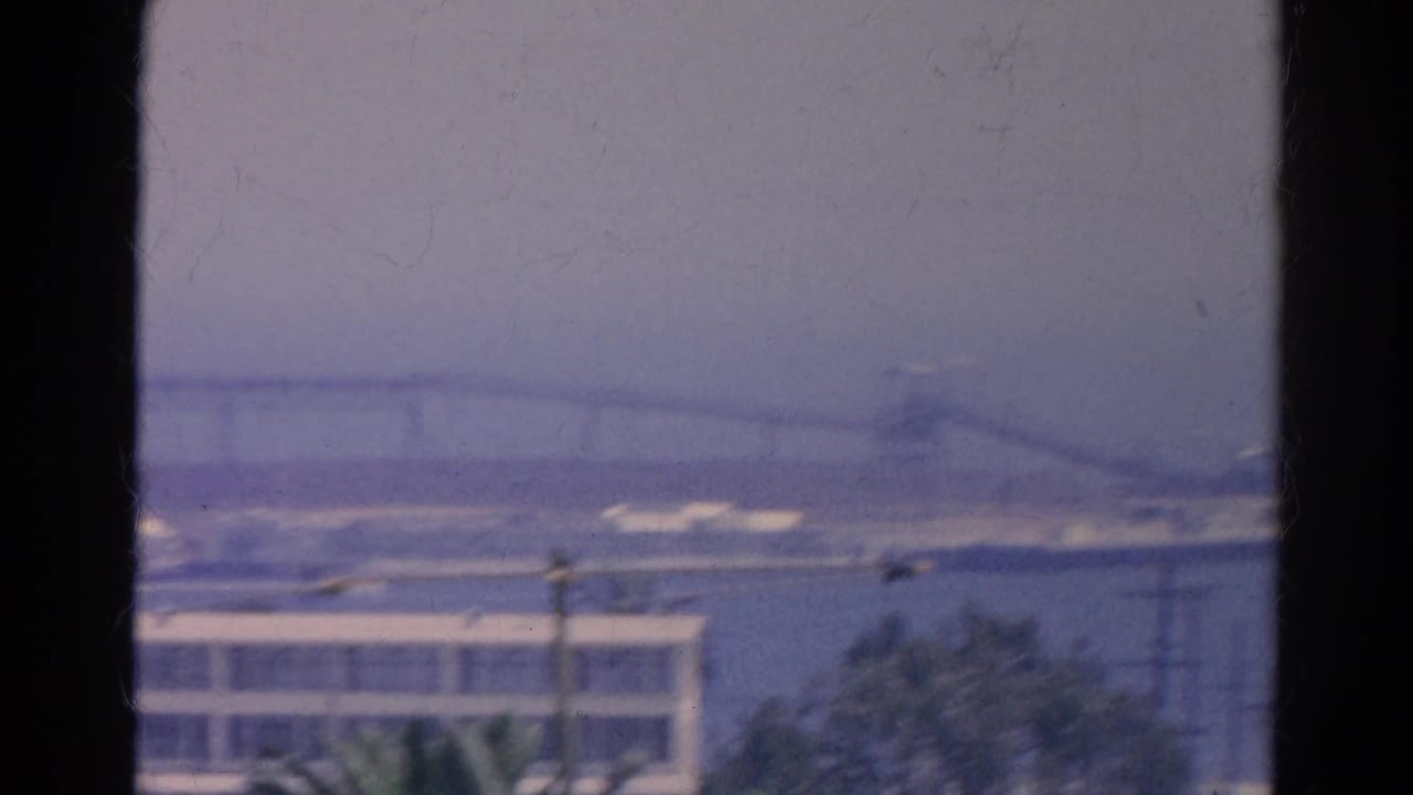 A hazy 1966 8mm view across water toward a long bridge, low buildings, and palms from a window.