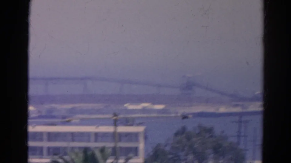 A hazy 1966 8mm view across water toward a long bridge, low buildings, and palms from a window.